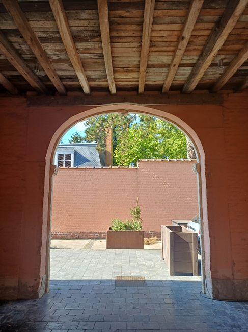 The image shows a view from inside a covered outdoor space with a wooden ceiling made of exposed beams and planks, leading through an arched opening to a paved courtyard. Outside, there is a large pink brick wall with a planter containing a small shrub or plant, and a tall tree beyond it, with bright green leaves indicating a sunny day. The courtyard features neatly laid paving stones and outdoor storage containers or compost bins positioned along the wall. In the background, parts of neighboring buildings and a chimney are visible, suggesting an urban setting. The scene appears to depict a garden or landscaped area in the process of maintenance or preparation, with clean, trimmed surfaces and a tidy arrangement, indicative of professional outdoor care by Gardeners Colliers Wood, as referenced in the page title about their work at Merton Abbey Mills courtyard.
