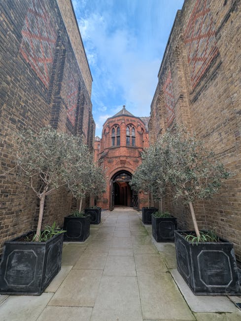 The image shows a view from inside a covered outdoor space with a wooden ceiling made of exposed beams and planks, leading through an arched opening to a paved courtyard. Outside, there is a large pink brick wall with a planter containing a small shrub or plant, and a tall tree beyond it, with bright green leaves indicating a sunny day. The courtyard features neatly laid paving stones and outdoor storage containers or compost bins positioned along the wall. In the background, parts of neighboring buildings and a chimney are visible, suggesting an urban setting. The scene appears to depict a garden or landscaped area in the process of maintenance or preparation, with clean, trimmed surfaces and a tidy arrangement, indicative of professional outdoor care by Gardeners Colliers Wood, as referenced in the page title about their work at Merton Abbey Mills courtyard.
